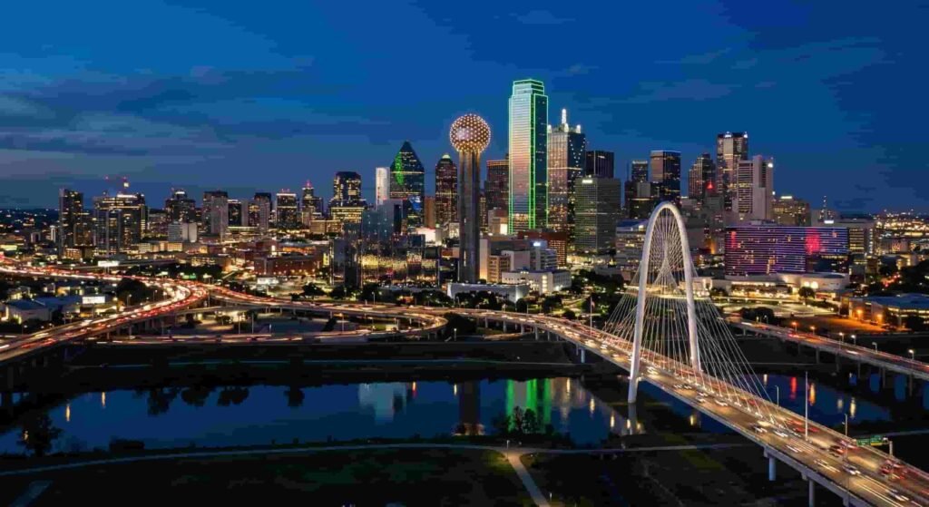 Night aerial photograph of the Dallas, Texas skyline, featuring the illuminated Reunion Tower, modern cable-stayed bridge, and busy highway traffic reflecting on the water below.